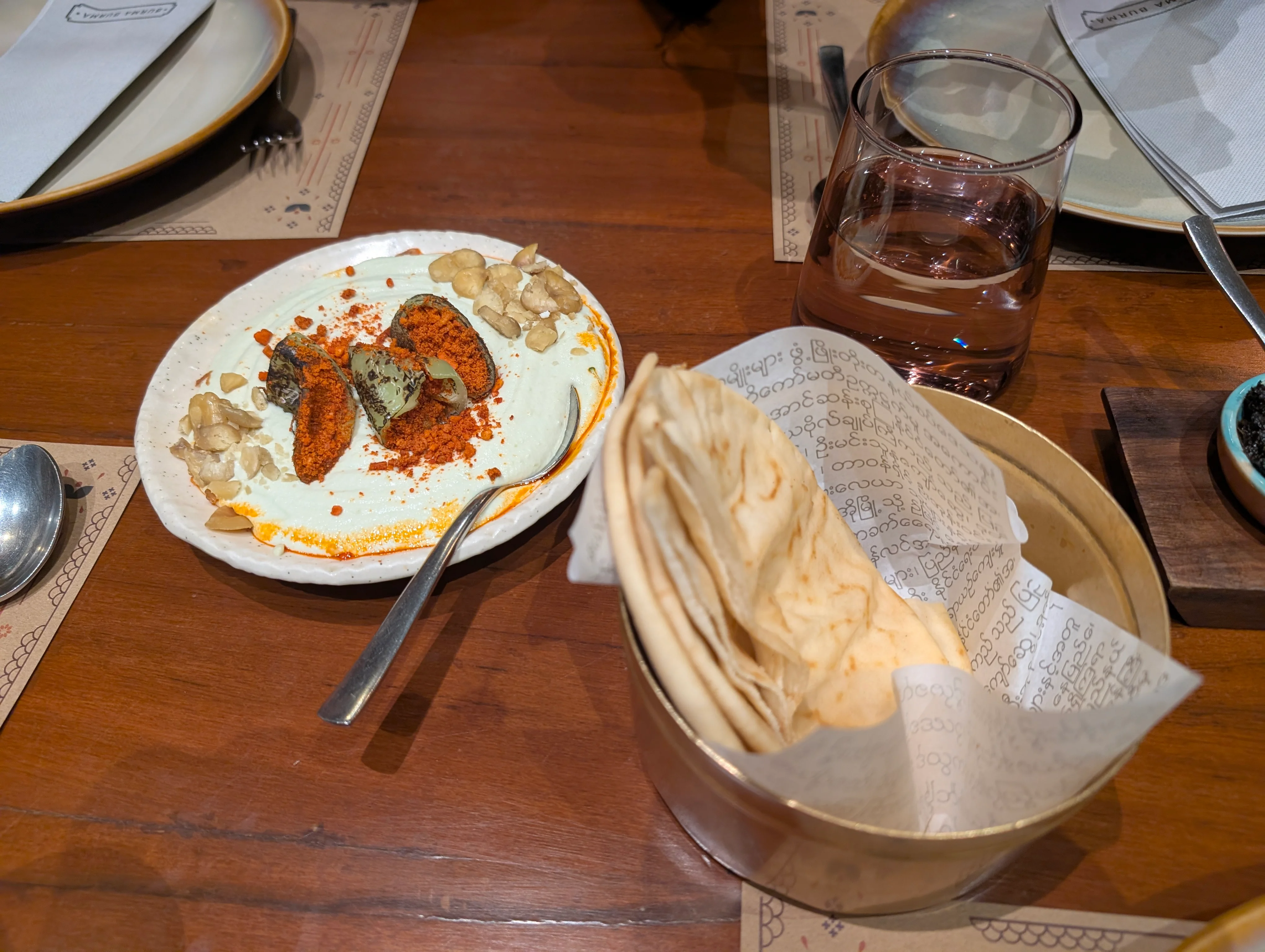 A basket of pita bread next to a plate of a bowl of hummus that has some big chillies and a red colored seasoning for garnish.