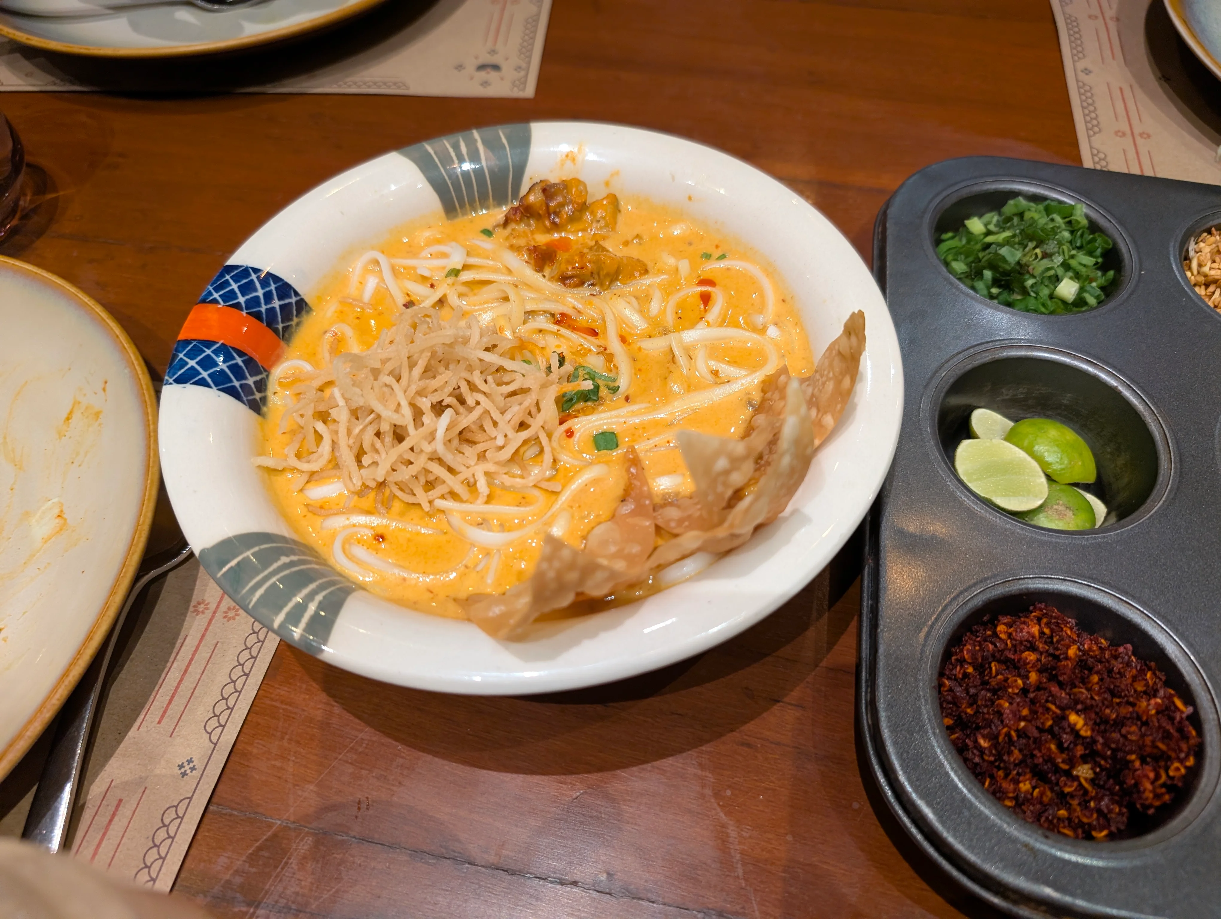 A bowl of khowsuey next to a condiments platter. The gravy is a bright yellowish orange in color and has regular flour noodles in it.
