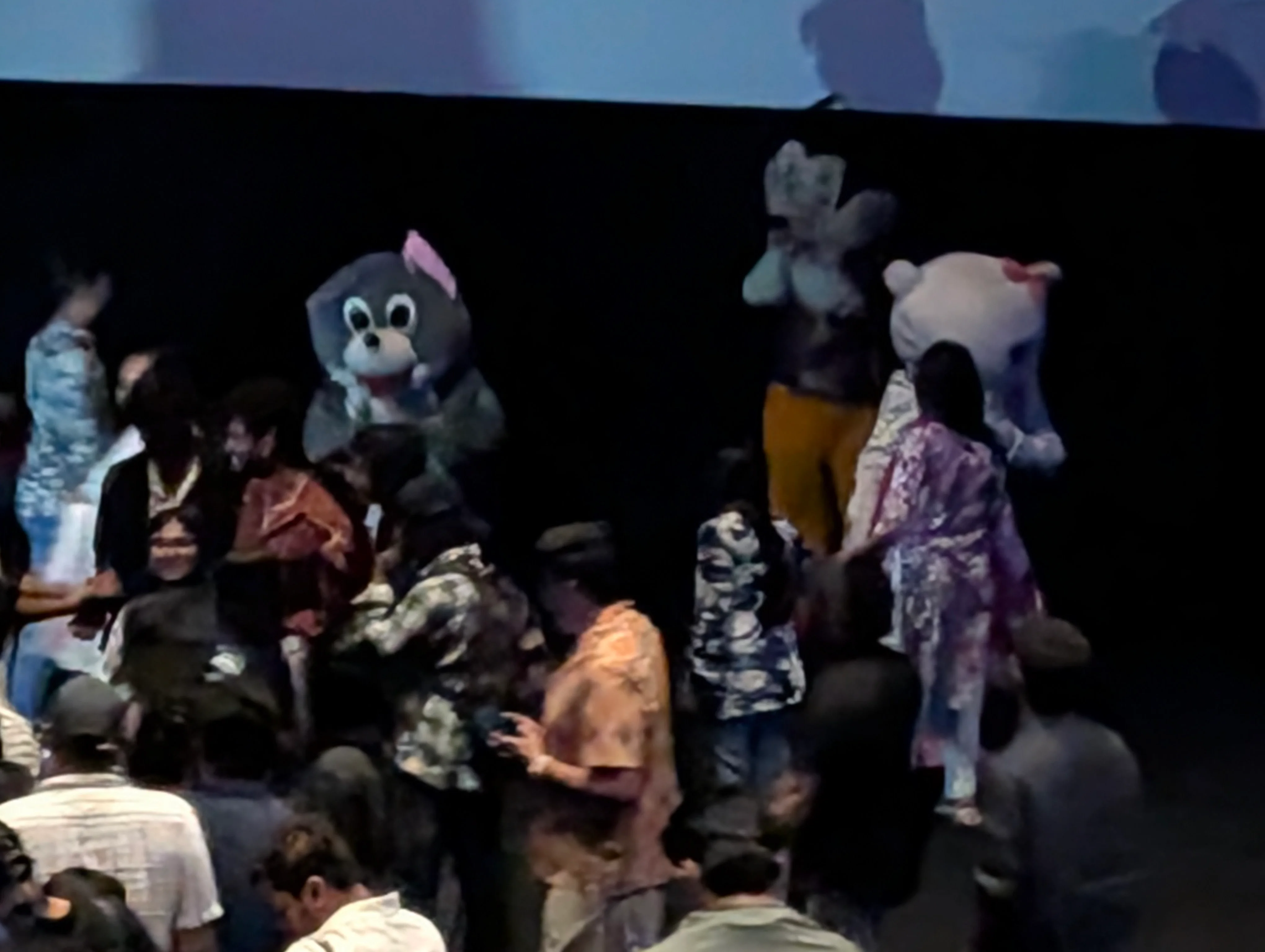 A slightly blurry zoomed in photo of the front of a movie theater, showing mascots in costumes: Hello Kitty, Mickey Mouse and a nondescript mouse I cannot identify immediately.